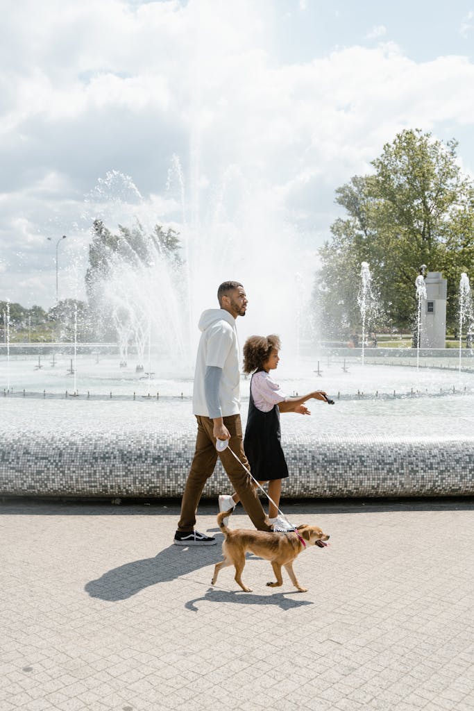 A father and daughter walk their dog by a large fountain in a park on a sunny day.