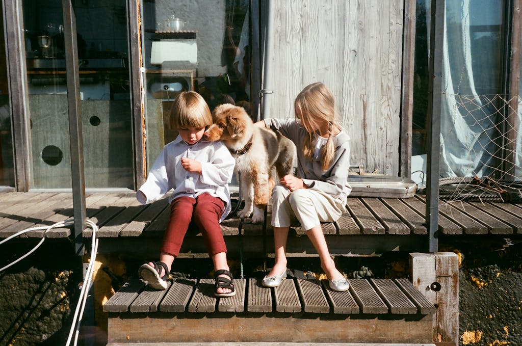 Two children sitting with a cute puppy on a wooden patio. Warm and playful outdoor scene.